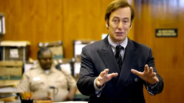 This image is a scene from the television show Better Call Saul. It shows the character Saul Goodman (real name Jimmy McGill), played by Bob Odenkirk, standing in a courtroom and speaking animatedly — likely addressing the judge or jury. He is dressed in a slightly flashy dark suit with a double-breasted jacket, a collared shirt, and a tie. His hands are raised mid-gesture, suggesting he's making an emphatic point or trying to persuade his audience.  In the background, a uniformed courtroom officer sits at a desk, looking relaxed, with shelves stacked full of paper files and folders behind him, emphasizing the formal and slightly cluttered court environment.  The tone of the scene reflects Saul Goodman's typical courtroom style — confident, theatrical, and slightly over-the-top — which is a hallmark of his character as a morally flexible lawyer who often bends the rules to win cases.