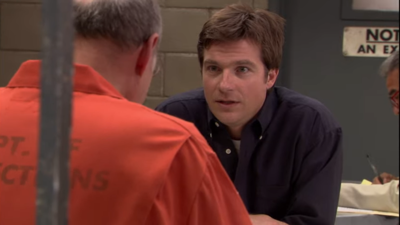 This image is a scene from the TV show Arrested Development. It takes place in a prison visiting room. On the left, an older man in an orange prison uniform, seen from behind, is speaking with a younger man across the table — his son, Michael Bluth, who is wearing a dark button-up shirt. They appear to be having a serious conversation.  This scene is known for a humorous moment where the father, George Bluth Sr., confidently tells his son, "They can’t charge a husband and wife for the same crime," implying it's a legal loophole — which is, of course, completely false and part of the show's satirical take on ignorance and corruption within the Bluth family.  The setting includes a cinderblock wall and a metal door in the background, reinforcing the prison environment. Another man is visible in the far right background, writing something, likely a prison guard or staff member. The mood of the scene combines seriousness with the show's signature dry humor.