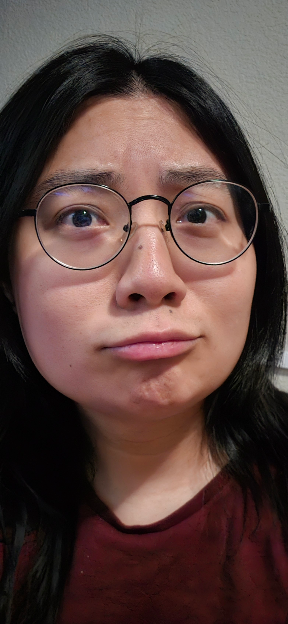 A close-up selfie of me making an exaggerated "trying not to cry" face: eyes wide, chin tucked, bottom lip pushed out in an obvious pout. Round wire glasses, dark red t-shirt, blank wall behind me.